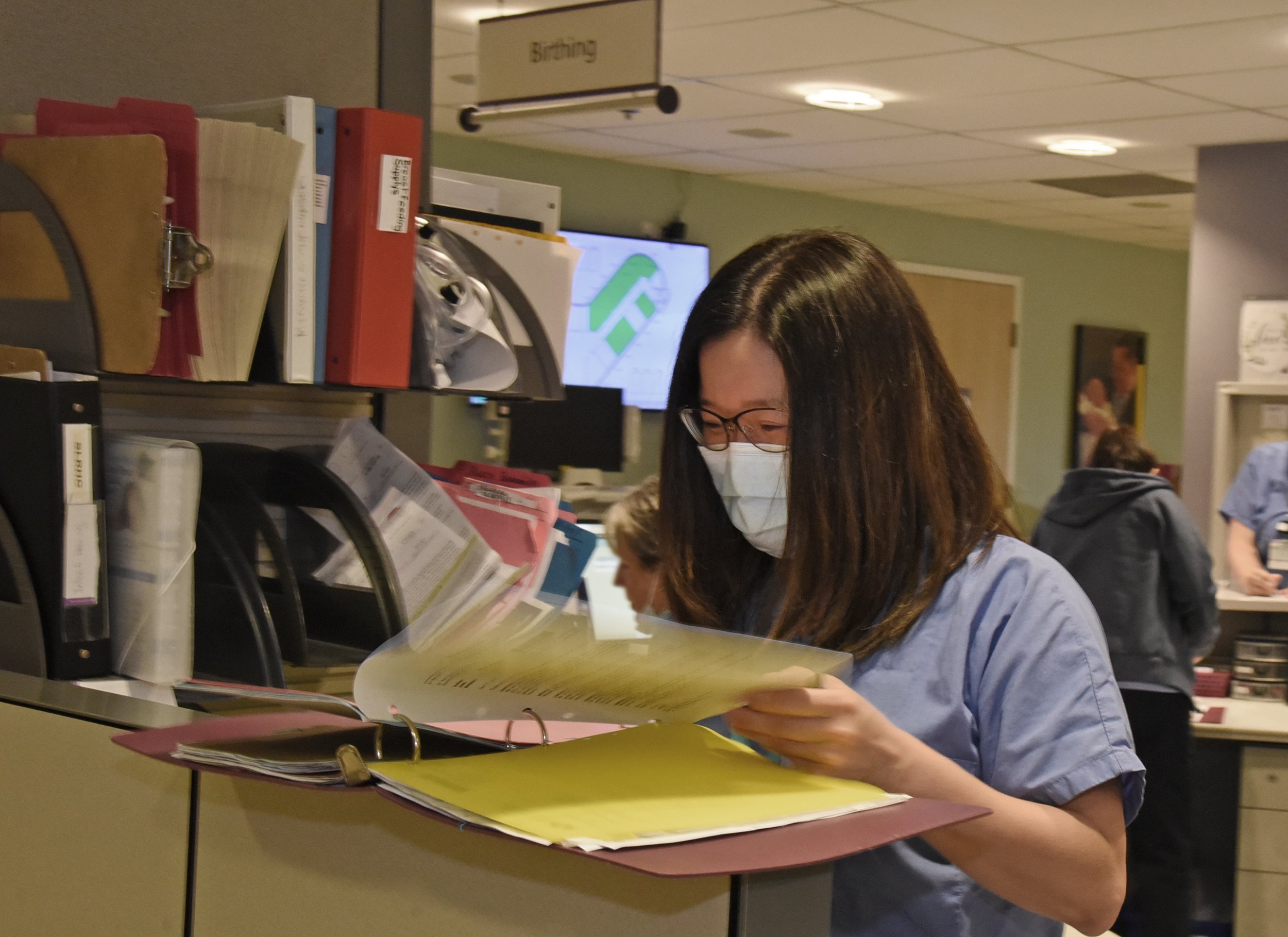 A woman wearing medical scrubs looks through a binder of documents